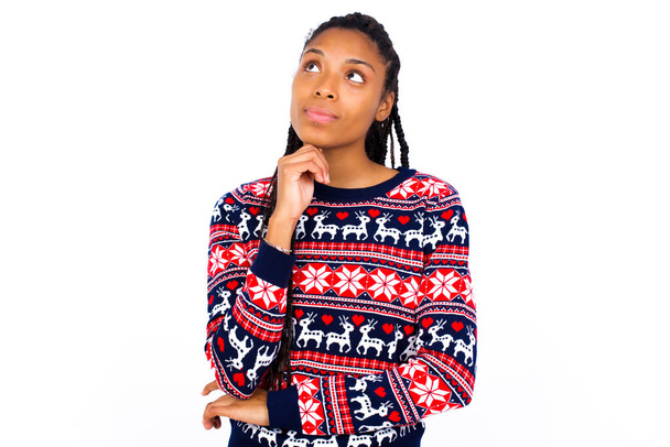 Wyrazy twarzy i emocje. Przemyślana Afroamerykanka ubrana w świąteczny sweter pod białą ścianą trzymająca go za rękę, mając wątpliwe spojrzenie. - Zdjęcie, obraz