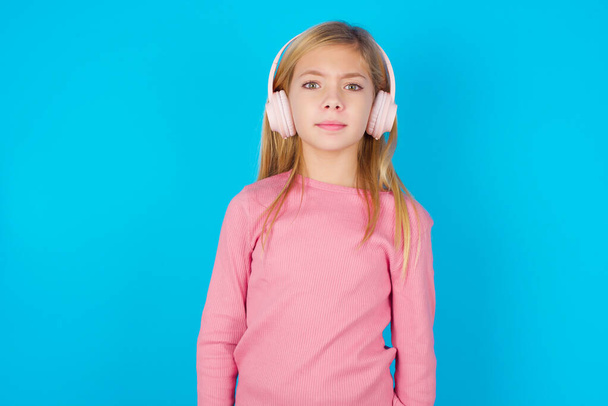 Serious displeased pretty teen girl looks puzzled at camera being angry wears stereo headphones listens music while walking at street - Photo, Image