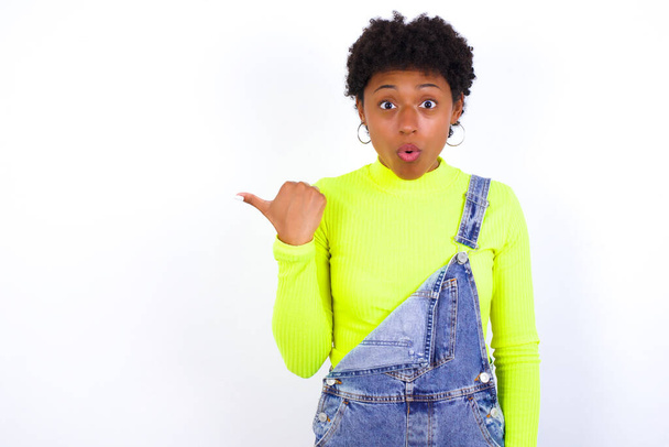 Zszokowana młoda Afroamerykanka z krótkimi włosami, nosząca dżinsy ogólnie przeciwko białym punktom ścian z kciukiem, coś wskazuje. Popatrz na to. Koncepcja reklamy. - Zdjęcie, obraz