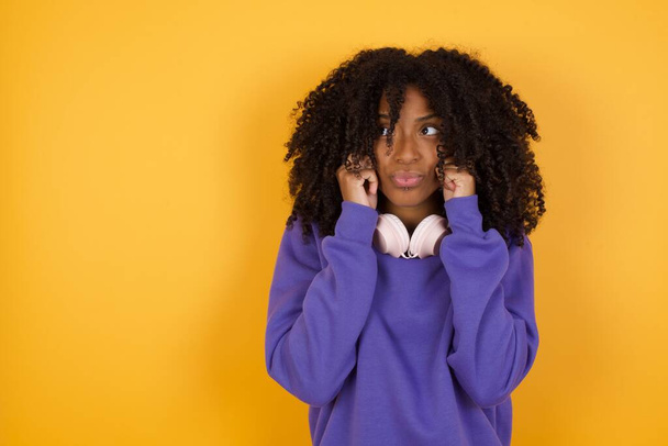 portrait de jeune femme afro-américaine expressive avec écouteurs sur fond jaune - Photo, image