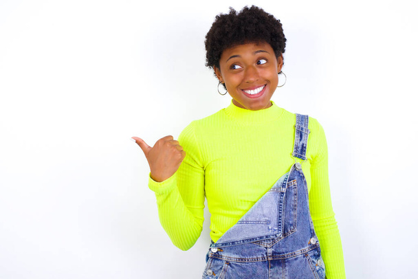 joven mujer afroamericana con el pelo corto con denim en general contra los puntos de la pared blanca de distancia y da consejos demuestra publicidad - Foto, imagen