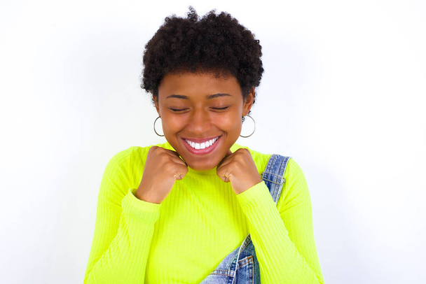mladá Afroameričanka s krátkými vlasy v džínsových kombinézách proti bílé stěně se radostně zazubí, představí si něco příjemného, kopírovací prostor. Příjemný koncept emocí. - Fotografie, Obrázek