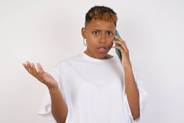 молода афроамериканка в білій футболці розмовляє по телефону над ізольованим тлом, напруженим рукою на обличчі, шокована соромом і несподіваним обличчям, розлючена і розчарована. Страх і засмучення за помилку. - Фото, зображення