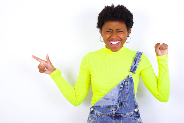 Χαρούμενη νεαρή Αφροαμερικανή γυναίκα με κοντά μαλλιά φορώντας τζιν συνολικά κατά λευκό τοίχο που δείχνει αντίγραφο χώρο αγγελία γιορτάζει την τύχη - Φωτογραφία, εικόνα