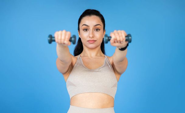 A healthy-looking young woman is training with dumbbells on a blue studio background. Shes got her sports outfit on and is staring at camera, a sign of her muscle and motivation - Φωτογραφία, εικόνα