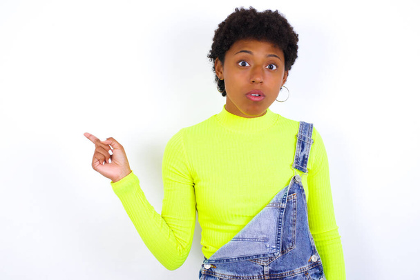 Emotive νεαρή Αφροαμερικανή γυναίκα με κοντά μαλλιά φορώντας τζιν συνολικά κατά λευκό τοίχο κρατά το σαγόνι έπεσε από το σοκ αποδεικνύει καταπληκτικό promo σημεία δεξιά στο κενό χώρο δείχνει μεγάλη πώληση. Διαφήμιση - Φωτογραφία, εικόνα