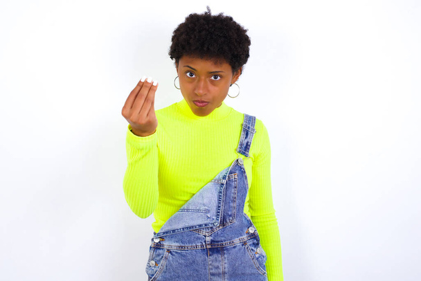 mladá Afroameričanka s krátkými vlasy v džínové kombinéze proti bílé stěně Dělat italské gesto s rukou a prsty sebejistý výraz - Fotografie, Obrázek