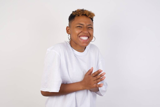 Νεαρή επιτυχημένη Αφρο-Αμερικανίδα που φοράει λευκό μπλουζάκι στέκεται στον λευκό τοίχο, αισθάνεται ευτυχισμένη, χαμογελά και χειροκροτεί, λέγοντας συγχαρητήρια με ένα χειροκρότημα. - Φωτογραφία, εικόνα