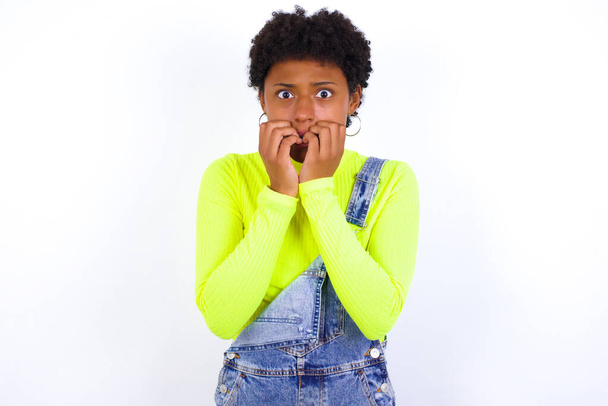 Άγχος - νεαρή Αφροαμερικανή γυναίκα με κοντά μαλλιά φορώντας τζιν συνολικά κατά λευκό τοίχο καλύπτει το στόμα του με τα χέρια φοβάται από κάτι ή κάποιος τρώει τα νύχια. - Φωτογραφία, εικόνα