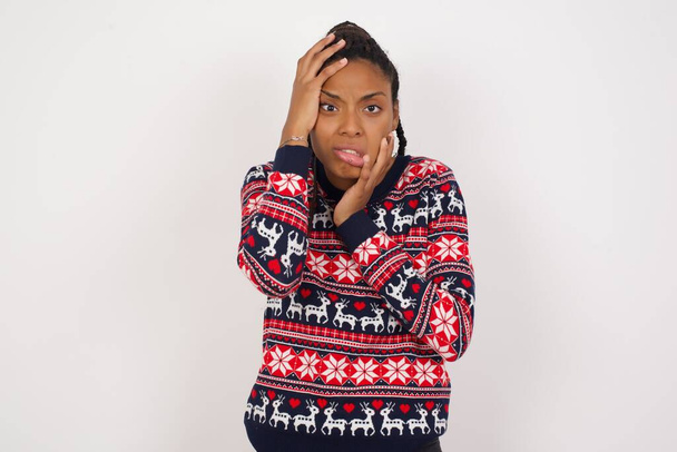 Junge düstere Afroamerikanerin trägt Weihnachtspullover vor weißer Wand, verdeckt Gesicht mit schmollenden und weinenden Händen, steht aufgebracht und deprimiert und klagt über Arbeitsplatzproblem. - Foto, Bild