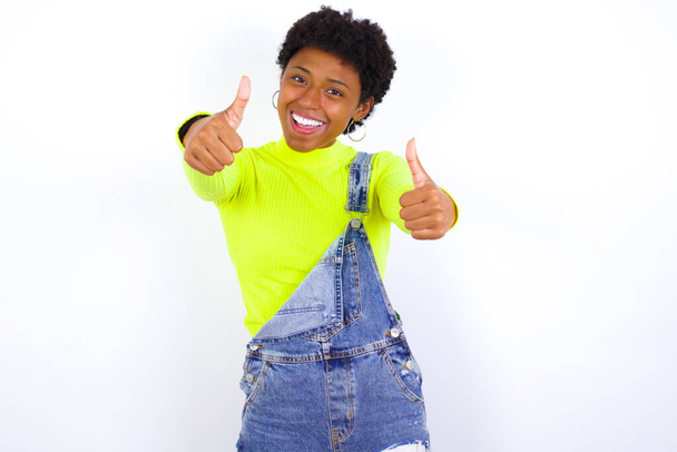 νεαρή Αφροαμερικανή γυναίκα με κοντά μαλλιά φορώντας τζιν συνολικά κατά λευκό τοίχο κάνοντας θετική χειρονομία με τους αντίχειρες επάνω χαμογελώντας και χαρούμενος για την επιτυχία. Κοιτάζοντας την κάμερα, νικήτρια χειρονομία. - Φωτογραφία, εικόνα