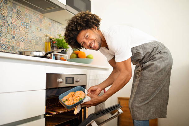 Schwarzer Mann schiebt fröhlich gewürzten Lachs in seiner heimischen Küche in den Ofen, trägt Schürze und lächelt in die Kamera. Guy kocht Fischmehl zum Abendessen, umarmt gesunde Ernährung - Foto, Bild