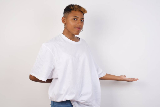 Porträt der schönen jungen Afroamerikanerin in weißem T-Shirt mit ausgestrecktem Arm in einer einladenden Geste vor grauem Hintergrund. - Foto, Bild