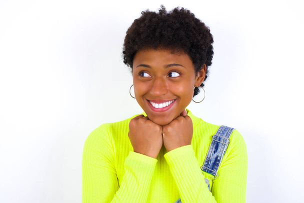 νεαρή Αφροαμερικανή γυναίκα με κοντά μαλλιά φορώντας τζιν συνολικά κατά λευκό τοίχο κρατά τα χέρια κάτω από το πηγούνι, στην ευχάριστη θέση να ακούσει εγκάρδια λόγια από ξένο - Φωτογραφία, εικόνα