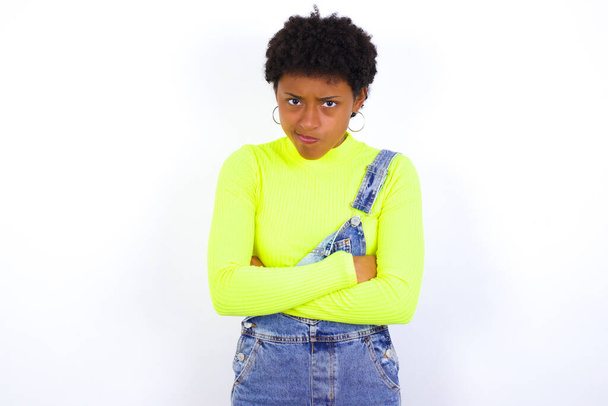 Foto de uma jovem afro-americana irritada com cabelo curto vestindo ganga geral contra os braços de cruzamento da parede branca. Olhando para a câmera com expressão decepcionada. - Foto, Imagem