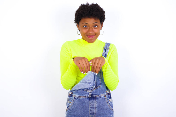 νεαρή Αφροαμερικανή γυναίκα με κοντά μαλλιά φορώντας τζιν συνολικά κατά λευκό τοίχο κάνει λαγουδάκι πόδια και φαίνεται με αθώα έκφραση παίζει με το μικρό παιδί της - Φωτογραφία, εικόνα