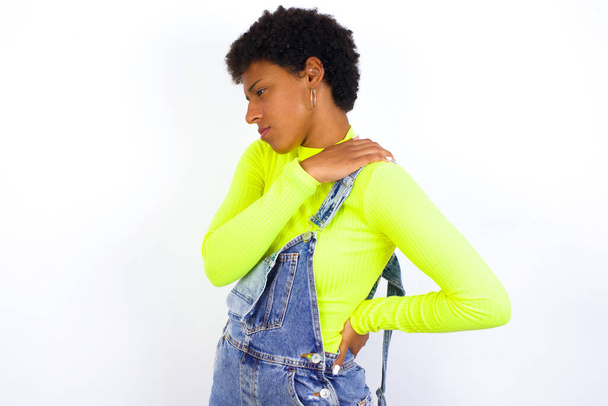 joven mujer afroamericana con pelo corto con denim en general contra la pared blanca tiene dolor de espalda - Foto, imagen