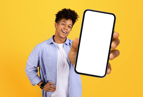 Happy young black man with curly hair holding smartphone with blank screen, ready for app promotion against vibrant yellow background - Photo, Image