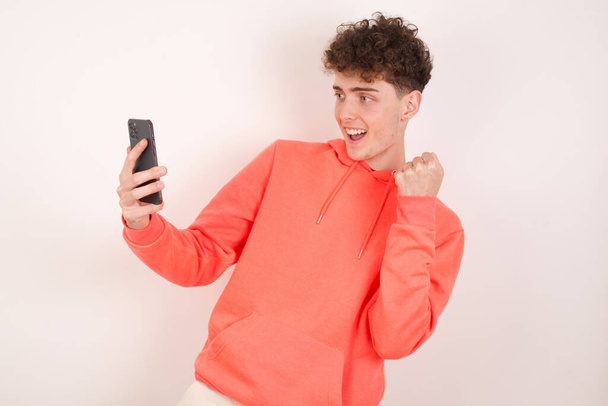 Ευτυχισμένος χαρούμενος όμορφος νεαρός άνδρας λαμβάνει καλά νέα μέσω e-mail και γιορτάζει την επιτυχία, ενώ στέκεται και κοιτάζοντας το κινητό τηλέφωνο. - Φωτογραφία, εικόνα