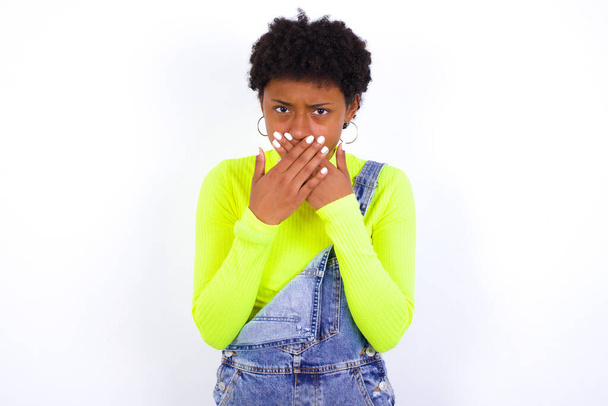 Mujer afligida afroamericana joven con el pelo corto que usa denim en general contra la pared blanca que cubre su boca con ambas palmas para evitar gritar, después de ver o escuchar algo malo. - Foto, imagen