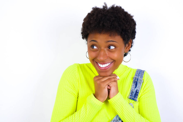 Šťastná mladá Afroameričanka s krátkými vlasy v džínové kombinéze na bílé stěně očekává něco úžasného, šťastně se dívá stranou, drží ruce u sebe, má šťastný výraz. - Fotografie, Obrázek