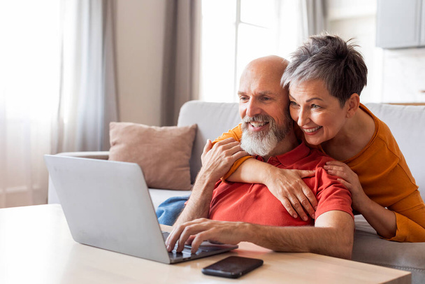 Улыбающиеся старшие супруги проводят время с ноутбуком в гостиной, счастливая пожилая пара с помощью компьютера дома, просмотр Интернета или покупки в Интернете, любящая жена обнимает мужа сзади - Фото, изображение