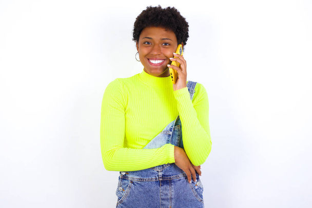 Portrét usměvavé mladé Afroameričanky s krátkými vlasy v džínsových kombinézách na bílé stěně, mluvící na mobilním telefonu. Obchodní, důvěra a komunikační koncepce. - Fotografie, Obrázek