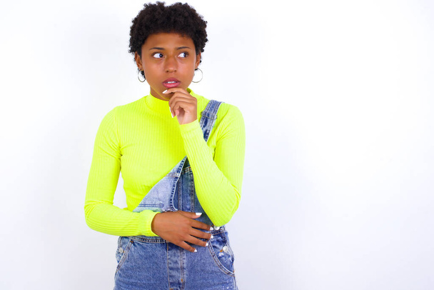 Знімок споглядальної молодої афроамериканської жінки з коротким волоссям, що носить джинсове загалом на білій стіні тримає руку під підборіддям, виглядає вдумливо вгору. - Фото, зображення