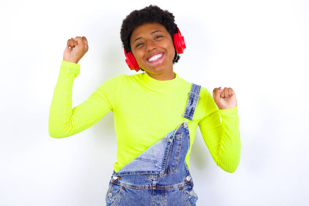Ξέγνοιαστη νεαρή Αφρο-Αμερικανίδα γυναίκα με κοντά μαλλιά που φοράει τζιν συνολικά ενάντια σε λευκό τοίχο με χαμόγελο στα πόδια αυξάνει τους χορούς αγκαλιά ξέγνοιαστες κινήσεις με ρυθμό της μουσικής ακούει μουσική από τη λίστα αναπαραγωγής μέσω ακουστικών - Φωτογραφία, εικόνα