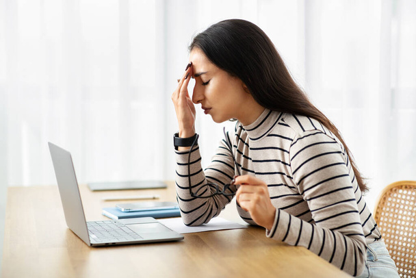 Mujer árabe joven triste trabajar en la computadora, sufren de migraña, problemas de salud, dolor y dolor de cabeza, sentirse mal en el interior de la sala de estar. Estrés, error, fecha límite en el ordenador portátil en casa - Foto, imagen