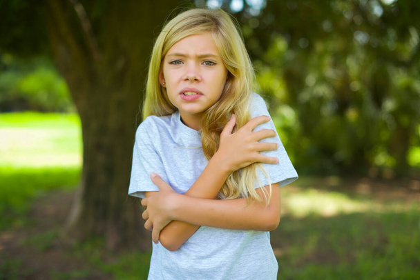 Πορτρέτο του καυκάσιου μικρού κοριτσιού που φοράει λευκό t-shirt και στέκεται έξω στο πάρκο αγκαλιάζοντας τον εαυτό της όταν αισθάνεται κρύο  - Φωτογραφία, εικόνα
