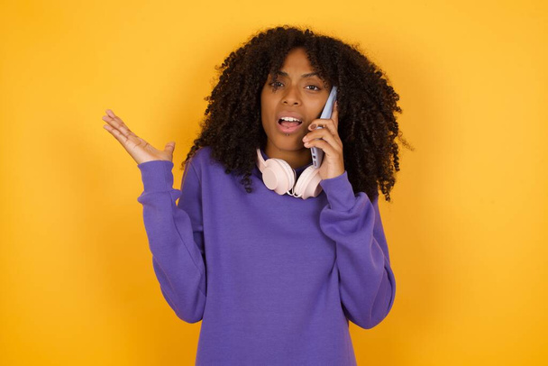 portrait of young expressive african american woman with headphones on yellow background talking by phone - Photo, Image