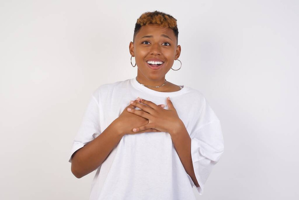 Ευτυχισμένη επιχείρηση Αφροαμερικανή γυναίκα φορώντας λευκό t-shirt στέκεται και χαμογελά απομονωμένη σε γκρι φόντο στούντιο έχει τα χέρια στο στήθος κοντά στην καρδιά. Νεαρή συναισθηματική γυναίκα. Ανθρώπινα συναισθήματα, έκφραση προσώπου. Πρόσθια όψη. - Φωτογραφία, εικόνα