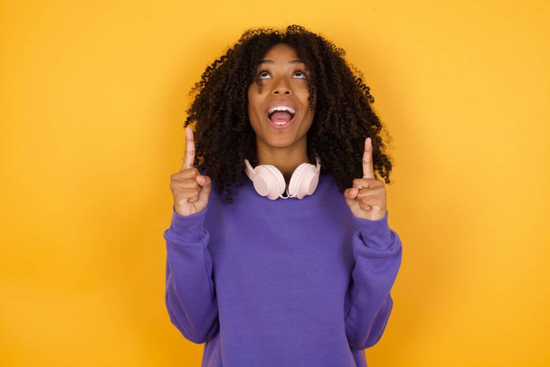 портрет молодої виразної афроамериканки з навушниками на жовтому тлі, що вказує вгору - Фото, зображення