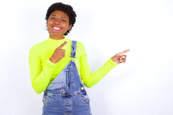 młoda Afroamerykanka z krótkimi włosami w dżinsach ogólnie na białej ścianie z pozytywnym wyrazem twarzy, wskazuje palcem wskazującym puste miejsce na tekst promocyjny lub reklamę. - Zdjęcie, obraz