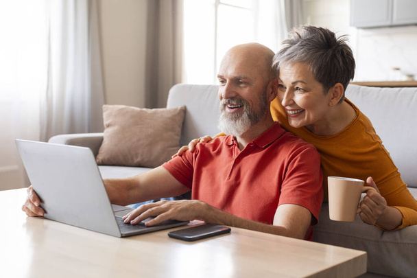Tech-Savvy Senioren. Gelukkig ouder paar met behulp van laptop samen thuis, Portret van vrolijke oudere echtgenoten surfen op internet op de computer, terwijl ontspannen in de woonkamer, genieten van moderne technologieën - Foto, afbeelding