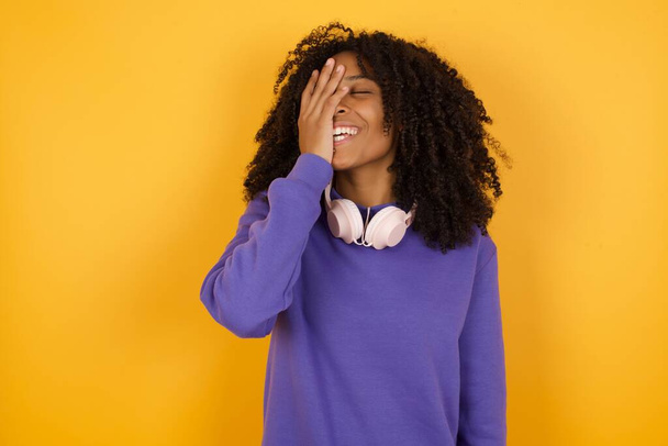 портрет молодої експресивної афроамериканки з навушниками на жовтому тлі сміється і покриває обличчя - Фото, зображення