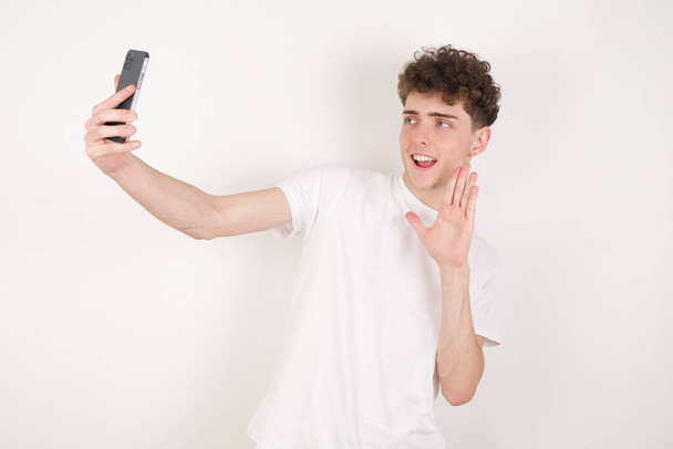 joven guapo sobre fondo blanco sostiene teléfono móvil moderno y hace que las ondas de videollamada palma en gesto de hola. Concepto de tecnología moderna - Foto, Imagen