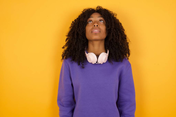 retrato de joven mujer afroamericana expresiva con auriculares sobre fondo amarillo mirando hacia arriba - Foto, Imagen