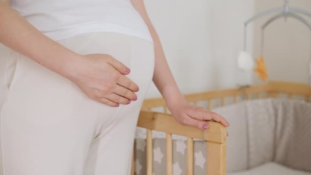 Pregnant woman stands near baby bed with hanging toys. Woman fingers trace contours of crib simultaneously caressing belly in anticipation of baby - Footage, Video