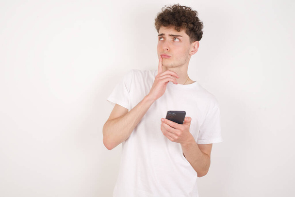 przystojny młody mężczyzna na białym tle myśleć głęboko o czymś, używa nowoczesnego telefonu komórkowego, stara się wymyślić dobrą wiadomość, trzyma palec wskazujący w pobliżu ust. - Zdjęcie, obraz