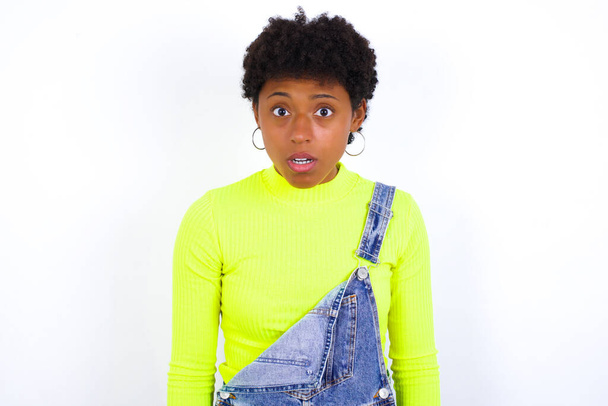 Emocionální atraktivní mladá Afroameričanka s krátkými vlasy, oblečená v džínovém kombinéze proti bílé stěně s otevřenými ústy, vyjadřuje velké překvapení a zděšení, zírá do kamery. Neočekávaná šokující zpráva a lidská reakce. - Fotografie, Obrázek