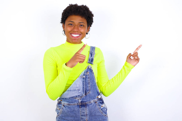 Joven mujer afroamericana positiva con el pelo corto usando denim en general contra la pared blanca con sonrisa radiante señalando con dos dedos y mirando en el espacio de copia vacío. Concepto de publicidad. - Foto, Imagen