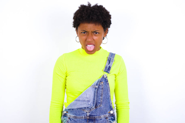 Körpersprache. Ekelhaft gestresste junge Afroamerikanerin mit kurzen Haaren trägt Jeans-Overall vor weißem Wand runzelndem Gesicht und demonstriert Abneigung gegen etwas. - Foto, Bild