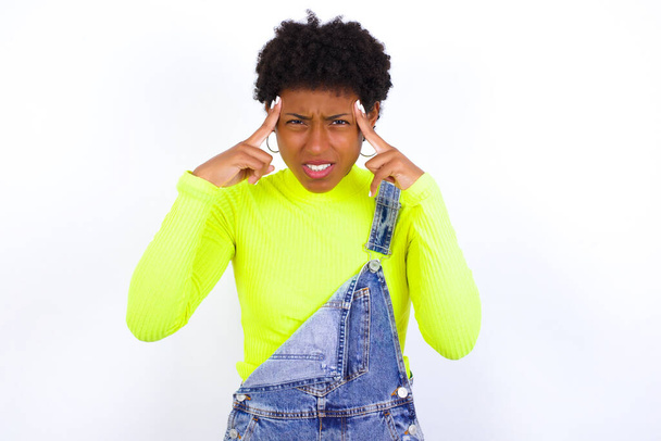 νεαρή Αφροαμερικανή γυναίκα με κοντά μαλλιά φορώντας τζιν συνολικά κατά λευκό τοίχο συγκεντρώνοντας σκληρά σε μια ιδέα με μια σοβαρή ματιά, σκέψης και με τα δύο δάχτυλα δείκτη δείχνει προς το μέτωπο. - Φωτογραφία, εικόνα