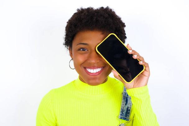 jeune afro-américaine aux cheveux courts portant globalement du denim contre un mur blanc tenant un smartphone moderne couvrant un œil tout en souriant - Photo, image