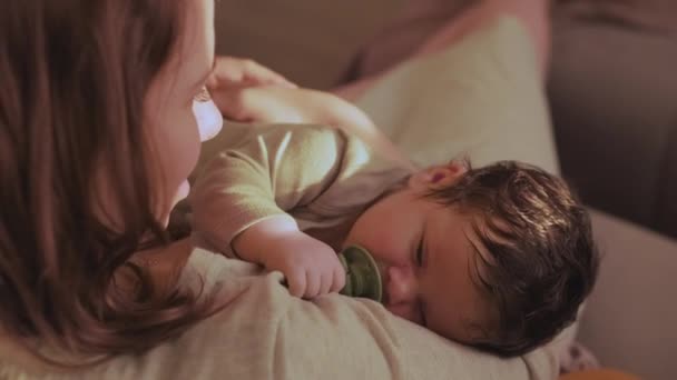 Moeder glimlacht terwijl ze de baby in haar armen houdt. zuigeling zuigt fopspeen en valt in slaap in zachte omhelzing van moeder onder zonlicht aanraken ouder gezicht - Video