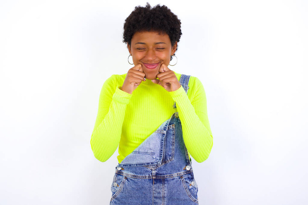 Χαρούμενη νεαρή Αφρο-Αμερικανίδα γυναίκα με κοντά μαλλιά που φοράει τζιν συνολικά ενάντια σε λευκό τοίχο με κλειστά μάτια κρατά τα χέρια κοντά στα μάγουλα και τα χαμόγελα τρυφερά φαντάζεται κάτι πολύ ευχάριστο - Φωτογραφία, εικόνα