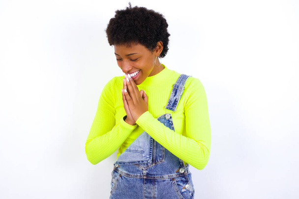 Ενθουσιασμένη νεαρή Αφρο-Αμερικανίδα με κοντά μαλλιά που φοράει τζιν συνολικά ενάντια στο λευκό τοίχο γελάει χαρούμενα και κρατά τις παλάμες πιέζονται μαζί ακούει κάτι αστείο - Φωτογραφία, εικόνα