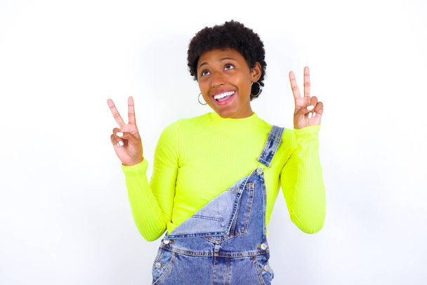 Απομονωμένη βολή της χαρούμενης νεαρής Αφροαμερικανής γυναίκας με κοντά μαλλιά φορώντας τζιν συνολικά κατά λευκό τοίχο κάνει την ειρήνη ή τη νίκη σημάδι με τα δύο χέρια, αισθάνεται δροσερό. - Φωτογραφία, εικόνα